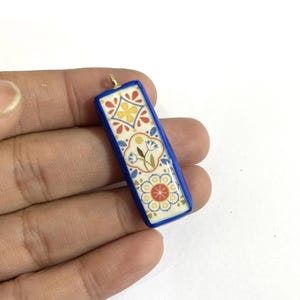 May include: A rectangular pendant with a blue border and a decorative floral pattern in red, yellow, and white. The pendant has a gold-coloured loop at the top for hanging. The design is reminiscent of traditional tile work.