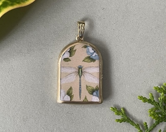 May include: A gold-colored pendant with an arched top. The pendant features a cream-colored background with a dragonfly illustration and floral accents in blue and white. The dragonfly has delicate wings and a slender body.