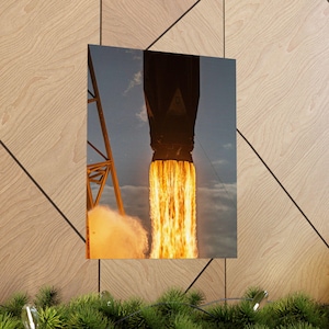May include: A framed print depicting a rocket launch. The image shows the rocket's exhaust plume in shades of orange and yellow against a blue sky. A yellow support structure is visible on the left side of the image.