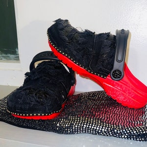May include: Two black and red clogs with a distressed denim fabric upper. The clogs have a red sole and gold studs around the edge.