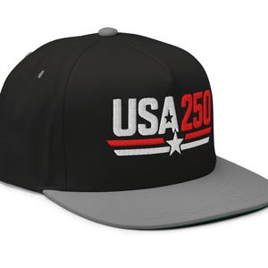 May include: A black baseball cap with a grey brim and a grey top button. The front features embroidered text that reads USA 250 in white and red, accompanied by a white star and two horizontal stripes in red and white positioned below the text.