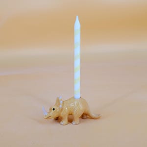May include: A ceramic triceratops-shaped candle holder in a light orange color, holding a white and yellow striped candle. The candle holder has a horn and three horns on its head. The candle is tall and thin.
