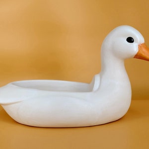 Op de afbeelding: Witte keramische eendenpot met een oranje snavel. De eend is gevormd als een pot met een uitgeholde body.