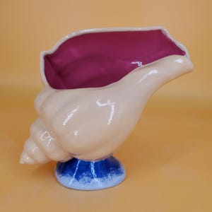 Puede incluir: Un macetero de cerámica con forma de concha marina con interior rosa, exterior blanco y base azul y blanca.