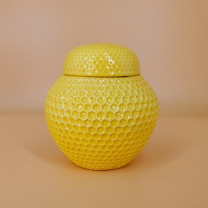 Puede incluir: Tarro de cerámica amarillo con tapa, adornado con un diseño de panal. El tarro tiene una forma redonda y abombada, mientras que la tapa es abovedada. El fondo es de color melocotón sólido.
