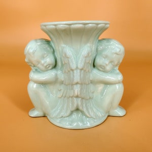 May include: A light green ceramic vase with two cherub figures on either side. The vase has a wide, round opening and a decorative design of wings on the front.