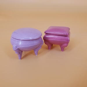 May include: Two ceramic trinket boxes. One is a light purple, round box with a lid and three curved legs. The other is a darker pink, square box with a lid and three curved legs. Both boxes are on a light orange background.