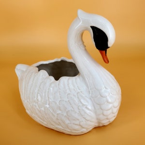 May include: White ceramic swan planter with a detailed feathered design. The swan's head is bent down and the beak is orange. The planter has a large opening for planting.