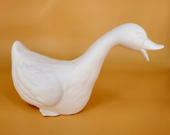 May include: White ceramic goose figurine with a long neck and a slightly open beak. The goose is standing on a flat surface.