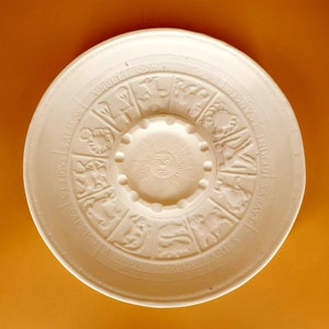 May include: White ceramic plate with a raised design of the zodiac signs and a sun in the center. The plate is circular and has a slightly raised rim.