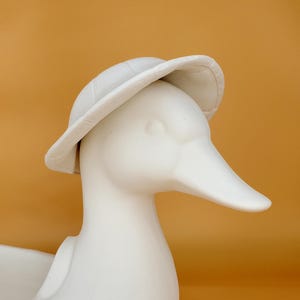 May include: White ceramic duck planter with a wide brimmed hat. The duck has a long beak and a rounded body. The planter is perfect for displaying small plants or flowers.