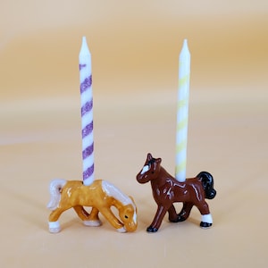 Puede incluir: Dos figuras de caballos de cerámica que sirven como portavelas. El caballo de la izquierda es de color marrón claro con crines y cola blancas, sosteniendo una vela blanca con rayas moradas brillantes. El caballo de la derecha es marrón oscuro con crines y cola negras, sosteniendo una vela blanca con rayas amarillas.