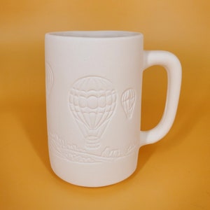 May include: White ceramic mug with a raised design of hot air balloons. The mug has a handle and is ready to be painted.