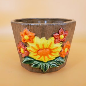 May include: A brown ceramic flower pot with a textured finish. The pot features a large yellow sunflower with orange and red accent flowers. The flowers are raised and have a glossy finish. The pot is empty and ready for planting.