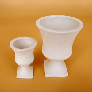 Może przedstawiać: Dwie białe ceramiczne urny w klasycznym stylu. Większa urna jest wyższa i szersza od mniejszej. Obie urny mają kwadratową podstawę i okrągłe ciało.