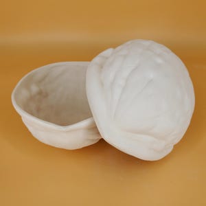 May include: A white ceramic bowl shaped like a walnut shell with a matching lid. The bowl is open and the lid is resting on top of the bowl.