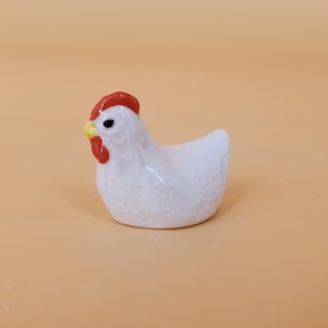 May include: A white ceramic chicken figurine with a red comb and yellow beak. The chicken is sitting on a light brown surface.
