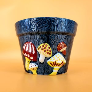 May include: A dark blue ceramic planter with a woodgrain pattern and mushroom designs. The mushrooms are painted in shades of red, yellow, and white.