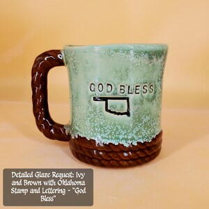 Könnte beinhalten: Eine Keramik-Tasse mit geflochtenem braunen Griff und Boden. Die Tasse hat eine hellgrüne Glasur mit weißen Sprenkeln und die Worte "GOD BLESS" über einer Umrisslinie von Oklahoma.