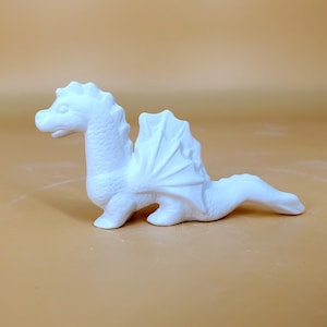 Puede incluir: Figurita de dragón de cerámica blanca con alas y cola larga. El dragón está en posición sentada con la cabeza girada hacia un lado.
