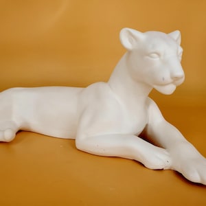 May include: A white ceramic sculpture of a reclining lioness. The feline figure is lying down with its front paws extended, and its head is raised. The sculpture has a smooth, matte finish and is set against a warm, golden-yellow background.