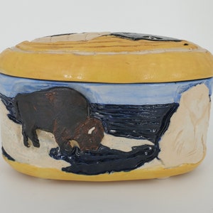 May include: A ceramic oval-shaped box with a yellow lid and a blue, white, and black scene on the bottom. The scene features a brown bison, a white figure, and a blue and black background.