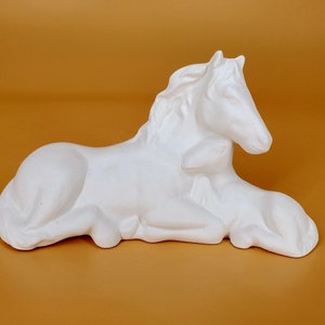 May include: White ceramic sculpture of a horse and foal lying down together. The horse has its head resting on the foal's back.