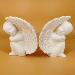 Könnte beinhalten: Zwei weiße Cherubim-Figuren, jede mit gefalteten Flügeln und einem ruhigen Ausdruck. Die Cherubim sitzen mit verschränkten Armen. Die Figuren bestehen aus einem glatten, matten Material.