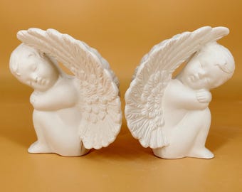 Może przedstawiać: Para białych, matowych figurek ceramicznych przedstawiających cherubiny w pozycji siedzącej, skulonej, z głowami opartymi na kolanach. Każda postać posiada duże, szczegółowe, pierzaste skrzydła rozciągające się w górę i na boki.