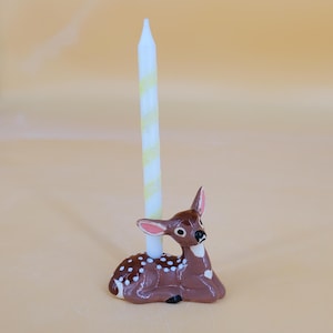 Puede incluir: Un ciervo de cerámica marrón con manchas blancas y una vela a rayas blancas y amarillas. El ciervo está tumbado, con la vela insertada en su espalda. El portavelas es un artículo decorativo.