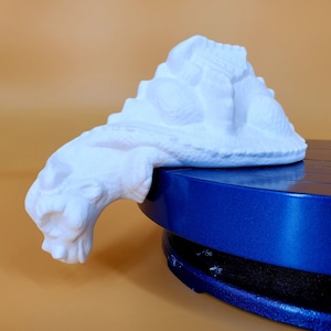 May include: White ceramic sculpture of a stylised mountain with a long, curved tail. The sculpture is resting on a blue turntable.