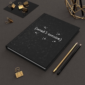 May include: A black notebook with a speckled texture and white text that reads "(sarah's version)" with small white stars. Two pencils, one gold and one black, are lying on a dark gray surface.