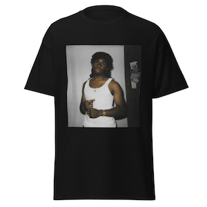 May include: Black and white photograph of a man wearing a white tank top and a gold chain. He is standing in a doorway with a white wall behind him.