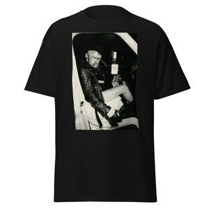 Op de afbeelding: Zwart-witfoto van een persoon die in een auto zit met een fles whisky in de hand. De persoon draagt een leren jack en een hoed.