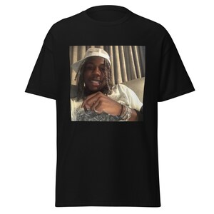 May include: A black t-shirt with a photo of a person wearing a white hat and smiling. The person has long dreadlocks and is wearing a white shirt.