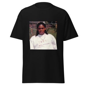 May include: A black t-shirt with a photo of a young person wearing a white Tommy Hilfiger sweatshirt. The person is standing in front of a green grassy area with a fence in the background.