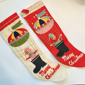 Puede incluir: Dos calcetines de Navidad vintage. Uno es rojo, el otro blanquecino. Cada uno tiene un diseño de fieltro de una marioneta en una bota negra, suspendida de un globo aerostático colorido. Las palabras "Merry Christmas" están escritas en la parte inferior de cada calcetín.