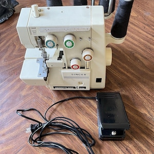 May include: A cream-coloured Singer Merrittlock 14U44 overlocker sewing machine with four black thread spools mounted on top. The machine features multiple tension dials with green, white, and red accents, and includes a black foot pedal with a power cord.