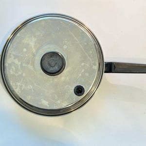Puede incluir: Una sartén de acero inoxidable con un mango negro y un asa lateral negra. La sartén tiene una tapa plateada con un pomo negro en el centro. La sartén está diseñada para cocinar.