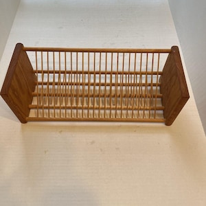 May include: A rectangular wooden record storage rack featuring two solid side panels and a series of thin, vertical dowels spaced evenly to create individual slots for organizing vinyl records. The structure is made of light brown wood.