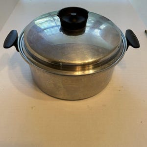 Puede incluir: Una olla redonda de acero inoxidable con tapa a juego. La olla cuenta con dos asas laterales de plástico negro y un pomo central de plástico negro en la tapa. La superficie metálica tiene un acabado pulido que refleja la luz.