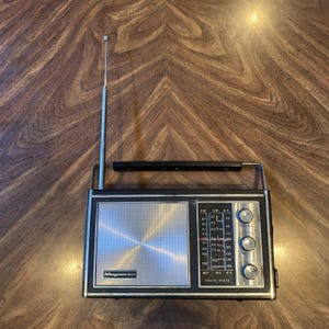 Könnte beinhalten: Ein tragbares Vintage-Radio von Magnavox mit schwarzem rechteckigem Gehäuse, silbernem Lautsprechergitter, vertikaler Teleskopantenne, schwarzem Tragegriff und einem Bedienfeld mit drei silbernen Drehknöpfen und einer Frequenzskala.