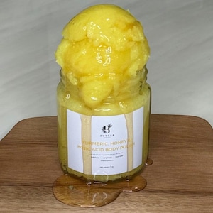 May include: A glass jar containing yellow body polish, with a large scoop of the same product on top. The jar has a white label with text: "Turmeric, Honey & Kojic Acid Body Polish". The product is on a wooden surface.