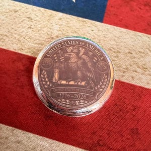 May include: A round, silver-colored coin with an eagle design and the text "UNITED STATES OF AMERICA 1776-2026". The coin is set against a backdrop of a red, white, and blue American flag.
