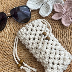 May include: A white crocheted sunglasses case with wooden beads and tassels. The case is on a woven wicker surface with two crocheted flowers in the background.