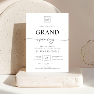 May include: A white invitation card with black text, announcing a grand opening. The card includes the words "GRAND opening" in elegant script, along with details about the event, such as the date, time, and location. The card is displayed on a neutral-toned surface.