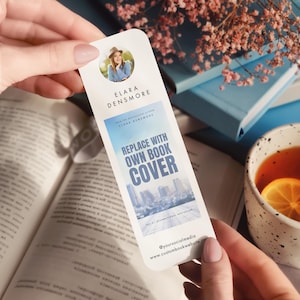 Peut inclure: Un marque-page blanc avec une photo d'une femme, le nom "ELARA DENSMORE", et le texte "REPLACE WITH OWN BOOK COVER". Le marque-page est tenu au-dessus d'un livre ouvert, avec une tasse de thé en arrière-plan.