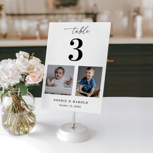 K&ouml;nnte beinhalten: Eine wei&szlig;e Tischnummerkarte mit der schwarzen Zahl 3, mit zwei Babyfotos. Auf der Karte steht "Sophie & Harold, 15. Oktober 20XX". Die Karte wird von einem silbernen St&auml;nder gehalten, und eine Vase mit Blumen befindet sich im Vordergrund.