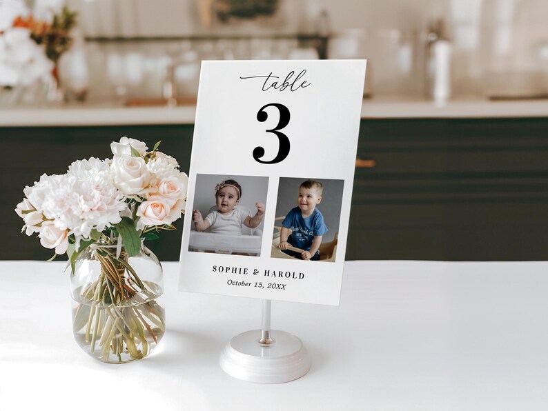 K&ouml;nnte beinhalten: Eine wei&szlig;e Tischnummerkarte mit der schwarzen Zahl 3, mit zwei Babyfotos. Auf der Karte steht "Sophie & Harold, 15. Oktober 20XX". Die Karte wird von einem silbernen St&auml;nder gehalten, und eine Vase mit Blumen befindet sich im Vordergrund.