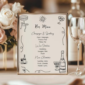 May include: A framed bar menu with a gold border, featuring black line art illustrations of champagne glasses, bottles, and decorative swirls. The menu lists champagne, wine, and non-alcoholic drink options.